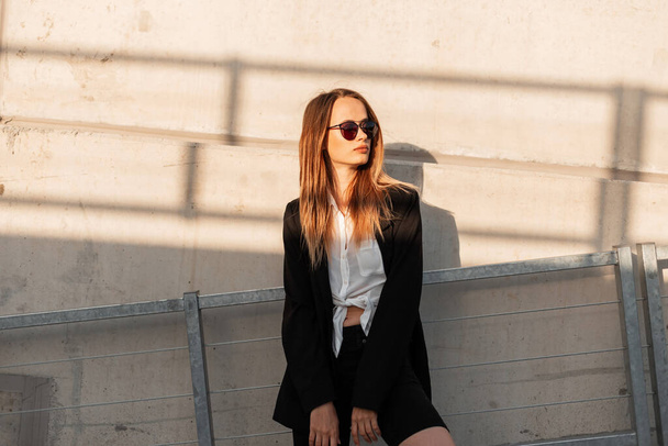 Moderne mooie jonge vrouw in vintage zonnebril in klassiek wit shirt in zwarte modieuze jas rust in de buurt van gebouw in de stad bij heldere zonsondergang. Aantrekkelijk cool meisje in de jeugd casual jurk buiten. - Foto, afbeelding