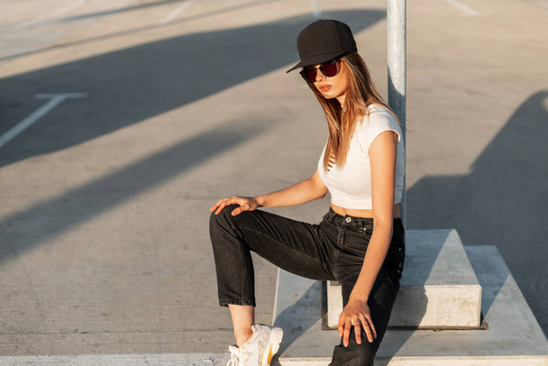 Amerykański stylowy hipster dziewczyna w moda czarny kapelusz w sexy biały top w modne dżinsy i trampki odpocząć na zewnątrz asfaltu w letni słoneczny dzień. Młoda miejska kobieta w luźnych ubraniach młodzieżowych na ulicy. - Zdjęcie, obraz