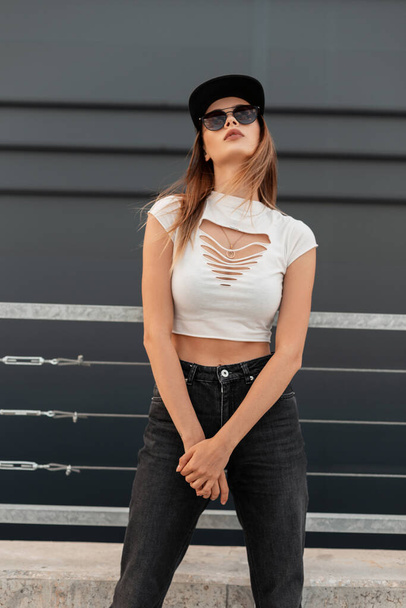 Μόδα ελκυστική αστική γυναίκα σε λευκό κομψό σκισμένο κορυφή σε τζιν σε δροσερό μοντέρνα γυαλιά ηλίου και μοντέρνο μπέιζμπολ μαύρο καπέλο σε γκρι φόντο σε εξωτερικούς χώρους στην πόλη. Casual καλοκαιρινή στολή νεολαίας. - Φωτογραφία, εικόνα