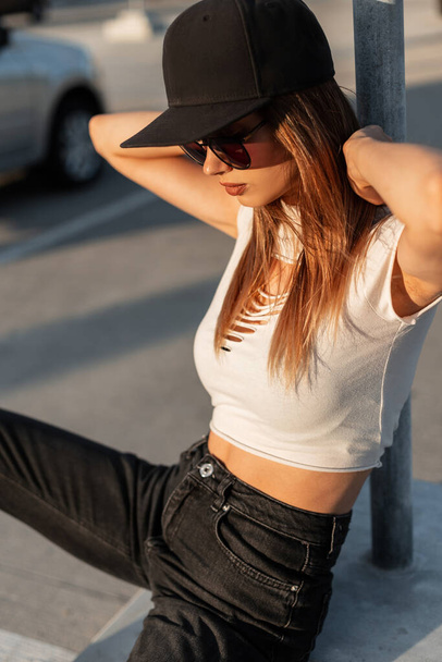 Urban νεαρή hipster κορίτσι με γυαλιά ηλίου σε μοντέρνα T-shirt σε μαύρο αμερικανικό καπέλο και τζιν στηρίζεται στον ήλιο στο πάρκινγκ της πόλης την ημέρα του καλοκαιριού. - Φωτογραφία, εικόνα