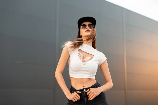 Sexy fashion model young woman in fashionable dark sunglasses in stylish youth cool clothes and american black cap poses near vintage wall in city at orange sunset. Casual summer style. - Фото, зображення