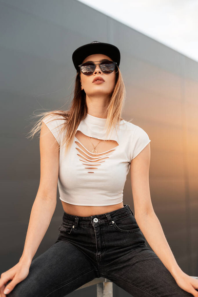 Slim sexy young hipster girl in fashionable dark sunglasses in stylish youth summer clothes from new collection and baseball black cap sit near vintage wall in city. Cool girl fashion model at sunset. - Photo, Image