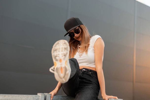 Attractive American woman in trendy black cap and sunglasses in stylish youth casual clothes shows leg in stylish sneakers to camera on gray background outdoors. Fashion city girl posing on at sunset. - Foto, imagen