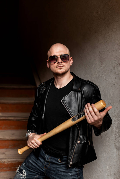 American brutal bald man with beard in fashion sunglasses in black vintage leather jacket in jeans stands with baseball bat indoors. Stylish cool bully guy poses near vintage stair. - Фото, изображение