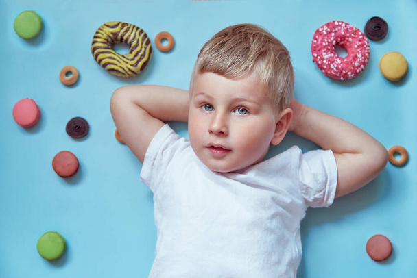 Lindo niño sonriente en donuts y macarrones franceses sobre fondo azul. Mock up camiseta blanca. Concepto del Día Nacional del Donut. Concepto de infancia feliz - Foto, Imagen