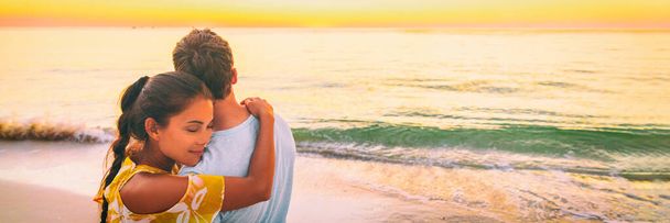 Couple in love hug banner panorama. Asian woman hugging embracing boyfriend relaxing on beach watching sunset - love and tenderness on travel summer holidays - Foto, afbeelding