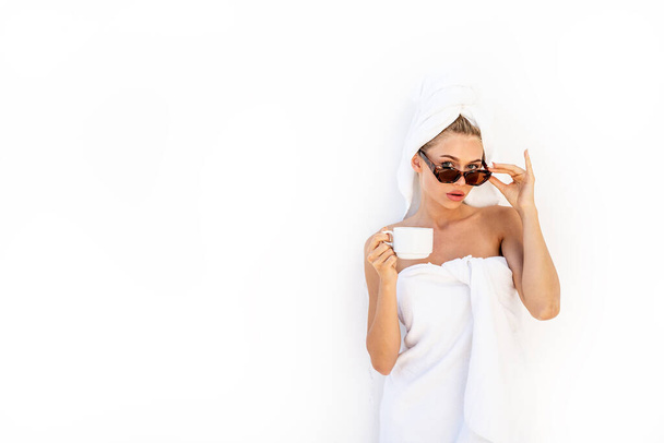 Jonge aantrekkelijke blanke vrouw na bad of douche genieten van zonnige dag. Meisje in witte handdoek met kopje koffie of thee met modieuze zonnebril. Luxe levensstijl. Rustig maar. Toerisme - Foto, afbeelding