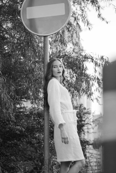 A confident girl walks down the street in a white home bathrobe. Fashion portrait. Idea for a photo shoot. Attracting attention. Stop road sign. Black and white photo  - Фото, зображення