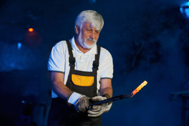 Aged blacksmith holding heated steel with forceps at forge - 写真・画像