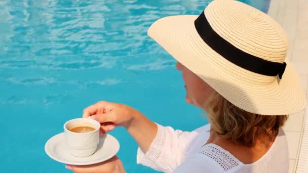 Primo piano di una donna di 50-55 anni con un cappello di paglia che beve da una tazza di caffè accanto a una piscina blu, lussuoso buongiorno, inizio giornata - Filmati, video