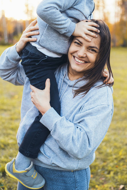 La mujer sostiene al niño en sus brazos mientras posan divirtiéndose juntos al aire libre. Retrato moderno. Una mujer joven y bonita. Cuidados familiares. - Foto, imagen