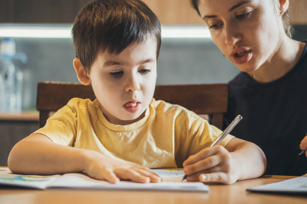 Niño haciendo su tarea con su madre sentada en el escritorio, en casa, que está escribiendo en el libro de texto. Desarrollo infantil. - Foto, imagen