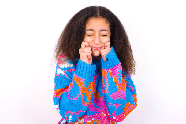 Pleased beautiful teen girl wearing colorful sweater standing against white background with closed eyes keeps hands near cheeks and smiles tenderly imagines something very pleasant - Photo, Image
