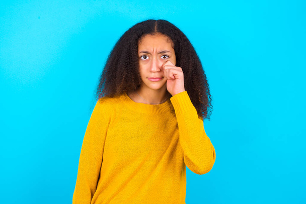 Zklamaný deprimovaný krásný dospívající dívka v žlutém svetru stojící proti modrému pozadí utírá slzy stojí ve stresu se zachmuřeným výrazem. Negativní emoce - Fotografie, Obrázek