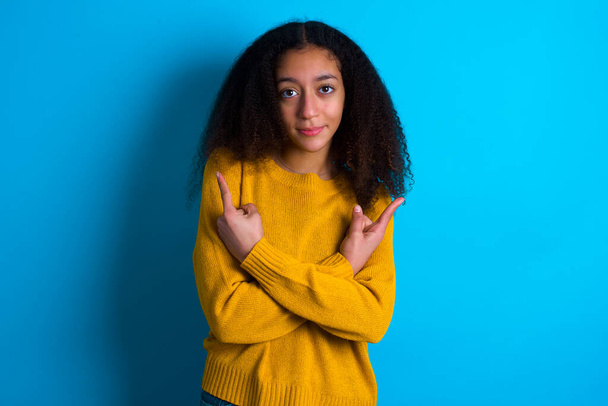beautiful teenager girl wearing yellow sweater standing against blue background crosses arms and points at different sides hesitates between two items or variants. Needs help with decision - Foto, afbeelding