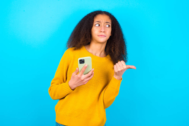 schönes Teenager-Mädchen in gelbem Pullover vor blauem Hintergrund zeigt Daumen weg und zeigt Leerzeichen zur Seite, hält Handy zum Senden von SMS. - Foto, Bild