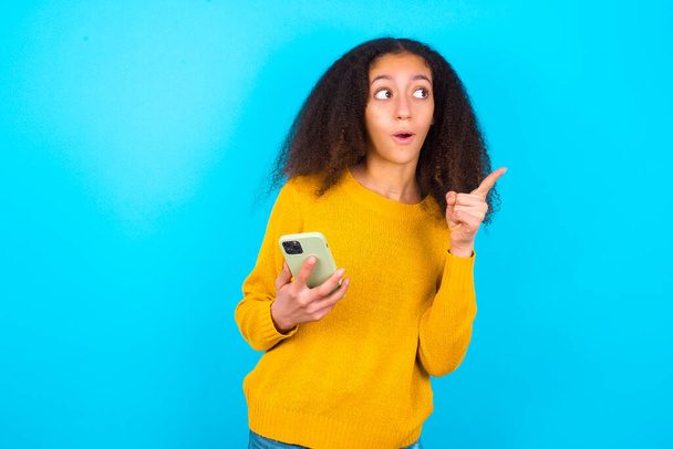 Stunned beautiful teenager girl wearing yellow sweater standing against blue background points sideways right copy space, recommends product, sees astonishing thing - Fotó, kép