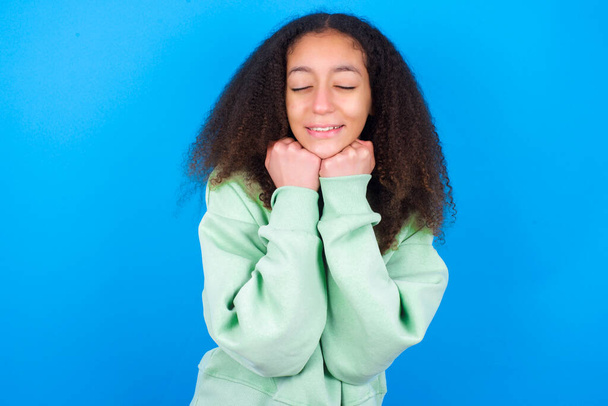 Веселая красивая девочка-подросток в зеленом свитере стоит на синем фоне имеет застенчивое удовлетворенное выражение лица, широко улыбается, показывает белые зубы, эмоции людей - Фото, изображение