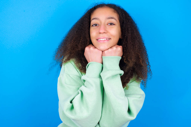 Zadowolona piękna nastolatka w zielonym swetrze stojąca na niebieskim tle dotyka podbródka obiema rękami, uśmiecha się przyjemnie, cieszy się dobrym dniem z kochankiem - Zdjęcie, obraz