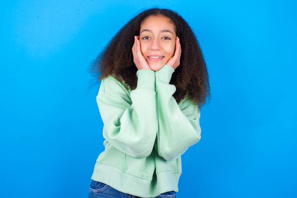 Boldog szép tinédzser lány visel zöld pulóver álló kék háttér érinti mindkét arcát finoman, gyengéd mosoly, mutatja fehér fogak, bámul pozitívan egyenesen a kamerába, - Fotó, kép