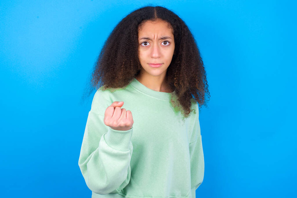 beautiful teenager girl wearing green sweater standing against blue background shows fist has annoyed face expression going to revenge or threaten someone makes serious look. I will show you who is boss - Photo, Image