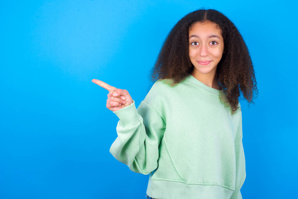Positiv schönes Teenager-Mädchen in grünem Pullover vor blauem Hintergrund mit zufriedenem Gesichtsausdruck zeigt oben rechts gutes Angebot an, auf Link klicken - Foto, Bild