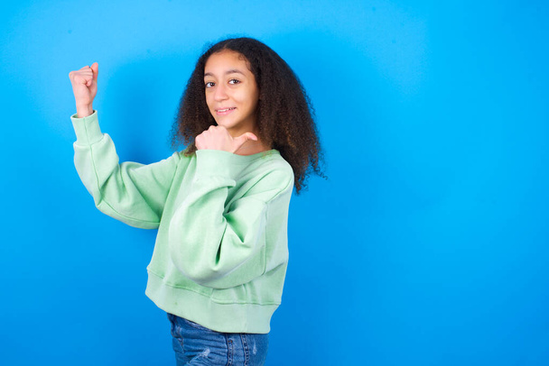 Hurá cool krásná teenager dívka na sobě zelený svetr stojící proti modrému pozadí bod zpět prázdné místo ruka pěst - Fotografie, Obrázek