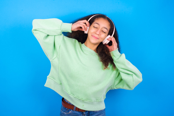 beautiful teenager girl wearing green sweater standing against blue background smiles broadly feels very glad listens favourite music track via wireless headphones closes eyes. - Fotografie, Obrázek
