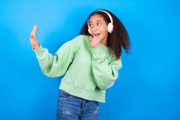 Happy beautiful teenager girl wearing green sweater standing against blue background sings favourite song keeps hand near mouth as if microphone wears wireless headphones, listens music - Photo, Image