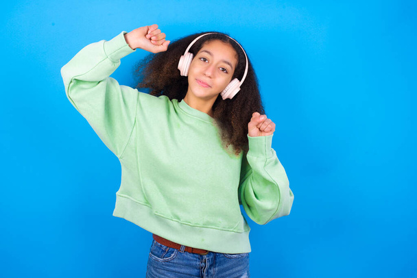 Carefree beautiful teenager girl wearing green sweater standing against blue background toothy smile raises arms dances carefree moves with rhythm of music listens music from playlist via headphones - Fotoğraf, Görsel