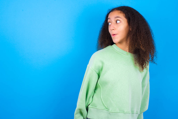 Megdöbbent gyönyörű tinédzser lány zöld pulóvert visel álló kék háttér néz üres tér nyitott szájjal sikoltozik: Ó, Istenem! Ezt nem hiszem el.. - Fotó, kép