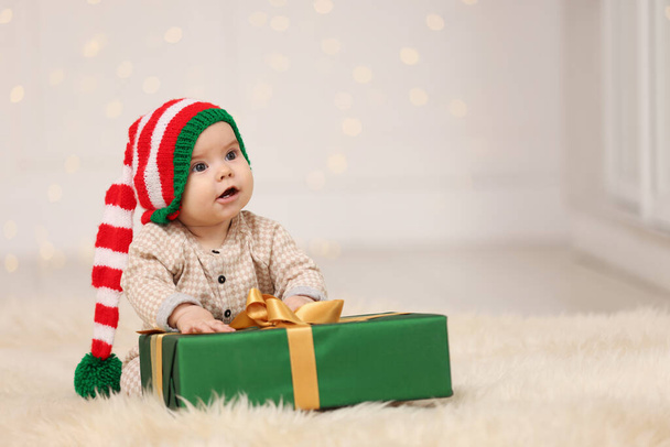 Cute baby w kapeluszu elfa z prezentem świątecznym na puszystym dywanie przed rozmytymi świątecznymi światłami, miejsce na tekst. Urlop zimowy - Zdjęcie, obraz