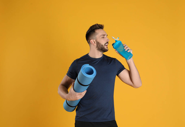 Handsome man with yoga mat drinking water on yellow background - Фото, зображення