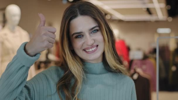 Junge schöne erfolgreiche kaukasische Mädchen Shopper Verbraucher Käufer stehen in Bekleidungsgeschäft und schauen in die Kamera lächelnd zeigt Daumen nach oben Zustimmung Empfehlung Zeichen der Zustimmung zustimmen Geste - Filmmaterial, Video