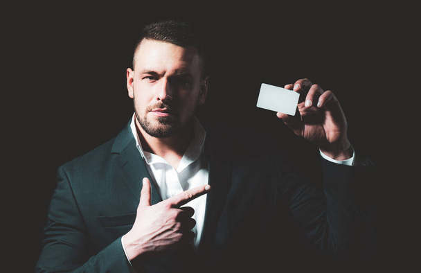 Portrait of a young man dressed in suit pointing finger at a credit card isolated over black background. Businessman with a credit-card. - Фото, зображення
