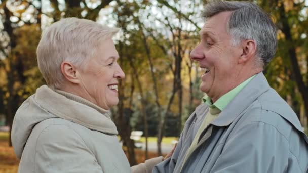 Side view loving old married couple hugging in autumn park smiling looking at each other happy pensioners laugh at good joke joyful elderly grandparents embrace outdoors harmony in family relationship - Footage, Video