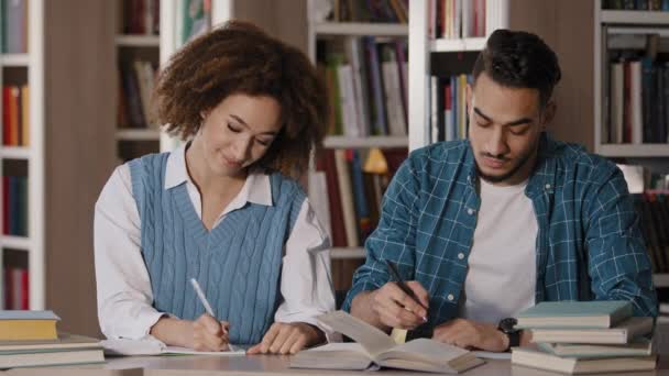 Δύο μαθητές, νεαρός και κορίτσι κάνουν εργασίες καθισμένοι στο γραφείο στη βιβλιοθήκη γράφοντας σημειώσεις ακούγοντας προσεκτικά τον λέκτορα στο μάθημα σε ομαδικές τάξεις σε συμμαθητές στο πανεπιστήμιο μιλώντας χαμογελαστά - Πλάνα, βίντεο
