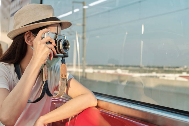 Krásná asijská turistka s obličejovou maskou sedí na červeném sedadle, cestuje vlakem, fotografuje, přepravuje se na předměstí, užívá si životního stylu cestujících po železnici, šťastnou dovolenou. - Fotografie, Obrázek