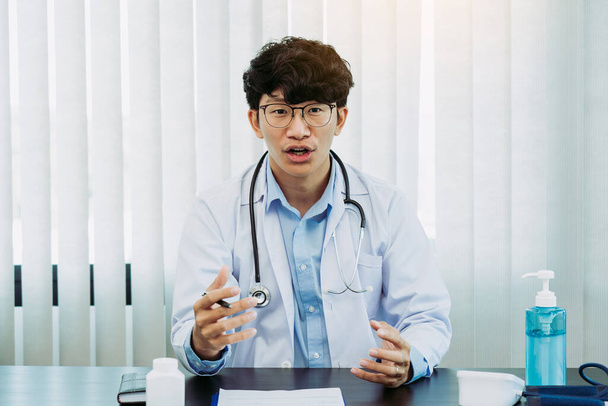 El médico asiático que usa anteojos y uniformes blancos con un estetoscopio proporciona asesoramiento en línea a los pacientes durante el brote de virus manteniendo una distancia social. - Foto, imagen