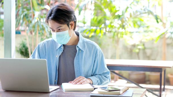 Young asian campus student man wearing protection mask working with laptop computer while online study in campus, coronavirus prevention in university, social distancing, online learning - Фото, изображение