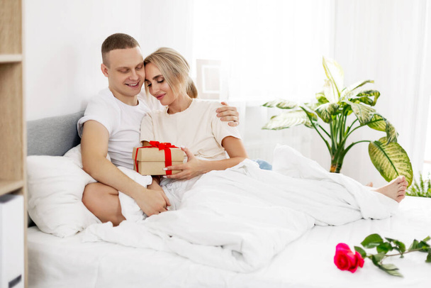 Ένα νεαρό κορίτσι με τον άντρα ή το αγόρι της ανοίγει δώρα το πρωί ξαπλωμένο στο κρεβάτι. Ημέρα του Αγίου Βαλεντίνου, επέτειος - Φωτογραφία, εικόνα