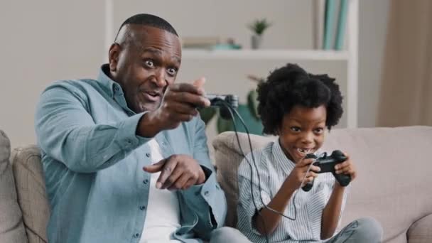 Зрілий афроамериканський батько з маленькою дочкою грає в відеоігри на консолі використовуйте джойстик контролер, сидячи на дивані насолоджуйтесь грою посмішка сміється тато запобігає дитячій дівчинці перемогти змагання
 - Кадри, відео