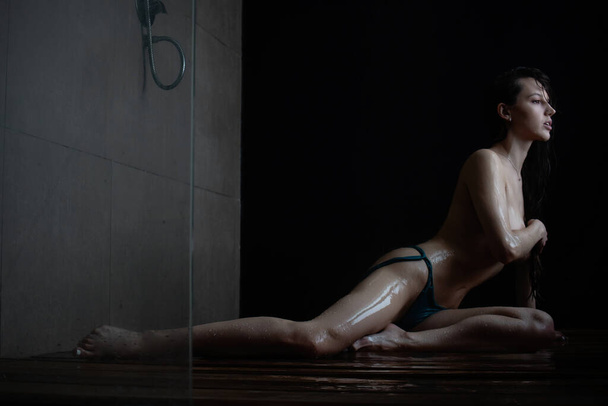 Side view full body of young slim sexy boso kobieta z ciemnymi włosami noszenie majtek bikini bez stanika siedzi na drewnianej podłodze pod prysznicem i pokrycie nagie piersi - Zdjęcie, obraz