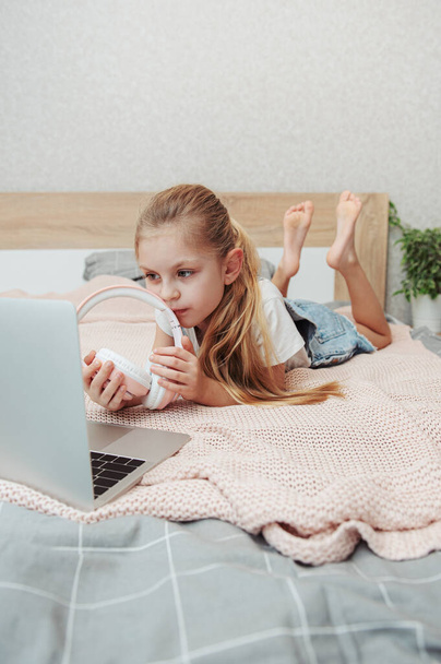 Посміхаючись щаслива маленька дівчинка, яка використовує ноутбук у бездротових навушниках у ліжку вдома
. - Фото, зображення