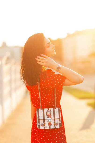 Muodikas kaunis nainen, jolla on kiharat hiukset punaisessa mekossa, jossa tyylikäs käsilaukku kävelee kadulla auringonlaskun aikaan. - Valokuva, kuva