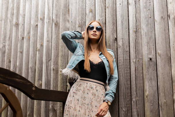 Pretty young girl model with vintage round sunglasses in a blue jeans jacket with black lace shirt and a skirt stands near a wooden wall - Foto, Bild