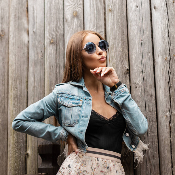 Elegante retrato de una hermosa mujer glamorosa con gafas de sol de moda en una chaqueta de mezclilla con lencería de encaje vintage y vestido posa cerca de una pared de madera. Chica enviando aire beso - Foto, imagen