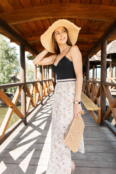 Fashionable pretty woman with a straw hat and a wicker bag in black lace lingerie with a vintage skirt is walking on a wooden terrace near the beach - Foto, imagen