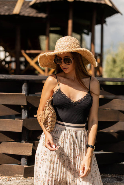 fashionable model woman in vintage round sunglasses with a straw hat with a woven handbag in stylish black lace lingerie and a skirt walks near the beach on summer holidays - Фото, зображення