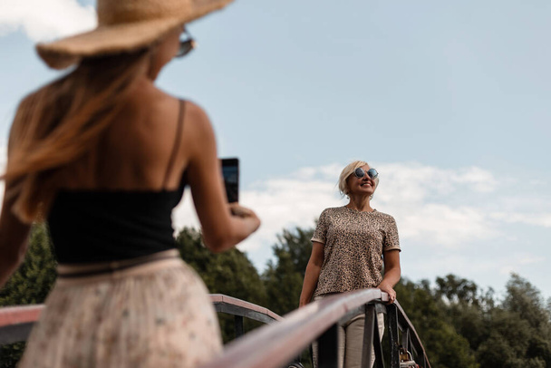 Hermosa hija de mujer joven en traje de verano de moda con un sombrero hace una foto en el teléfono de una madre anciana en una elegante camiseta estampada de leopardo al aire libre en fin de semana soleado - Foto, Imagen